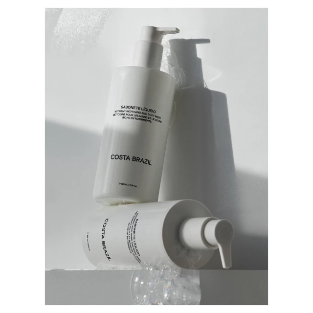 Two Costa Brazil Sabonete Liquido bottles with pump dispensers, one lying down and one standing, against a bright white background with bubbles.