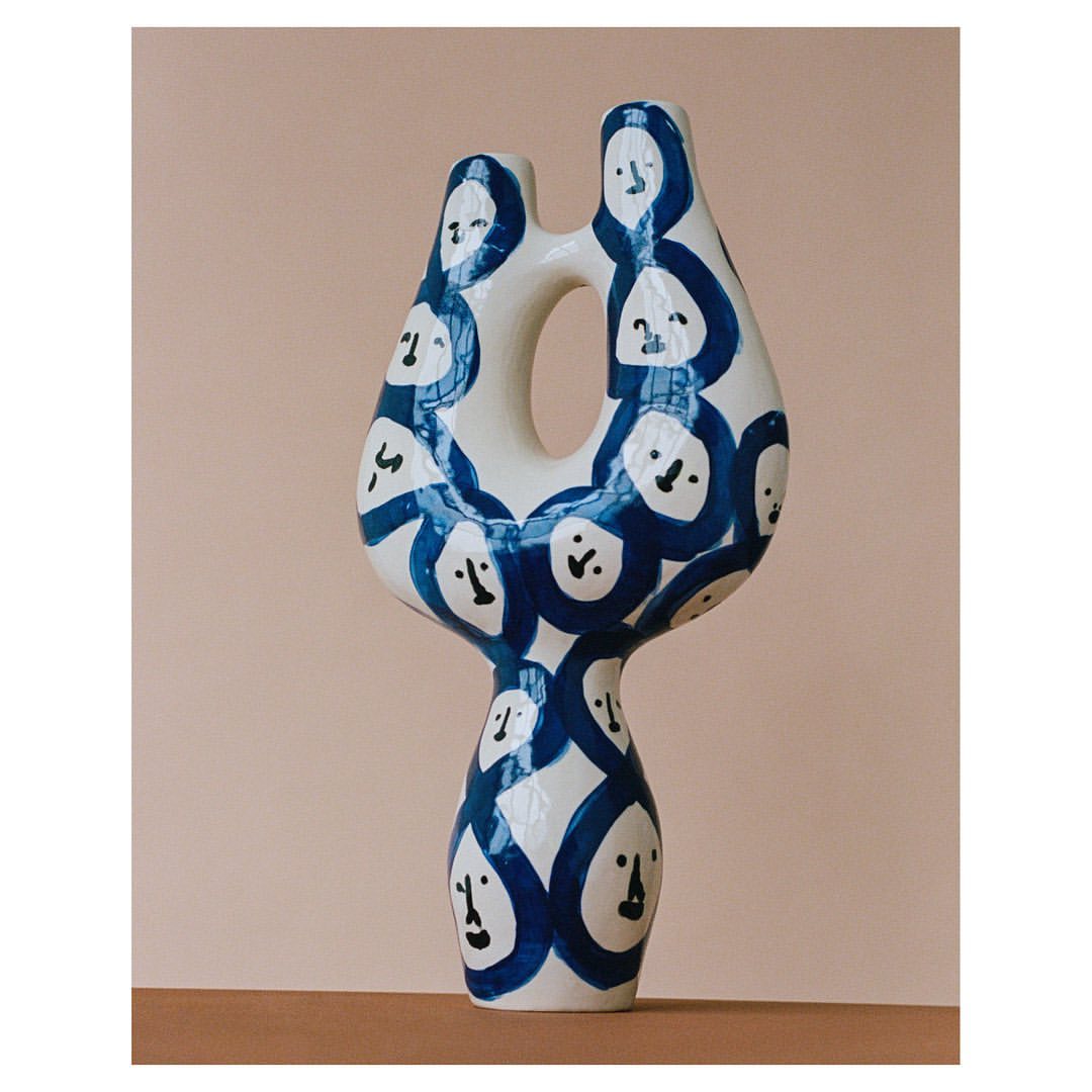 Modern white vase with abstract blue face illustrations, against a tan background with natural light casting soft shadows.