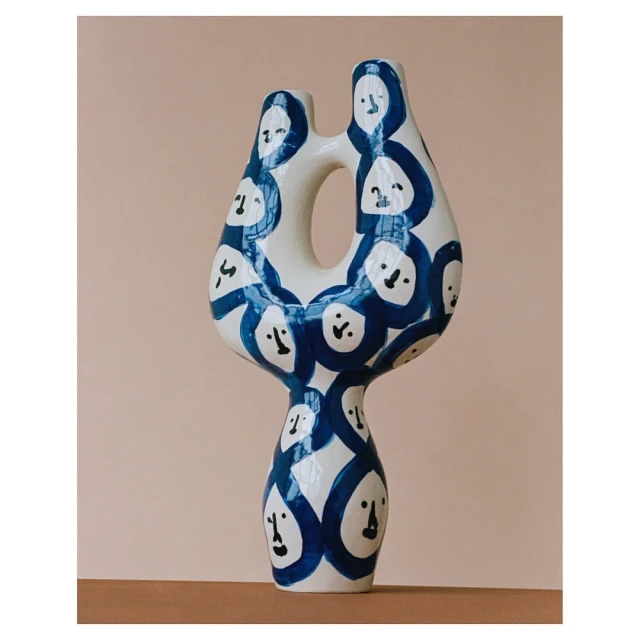 Modern white vase with abstract blue face illustrations, against a tan background with natural light casting soft shadows. 