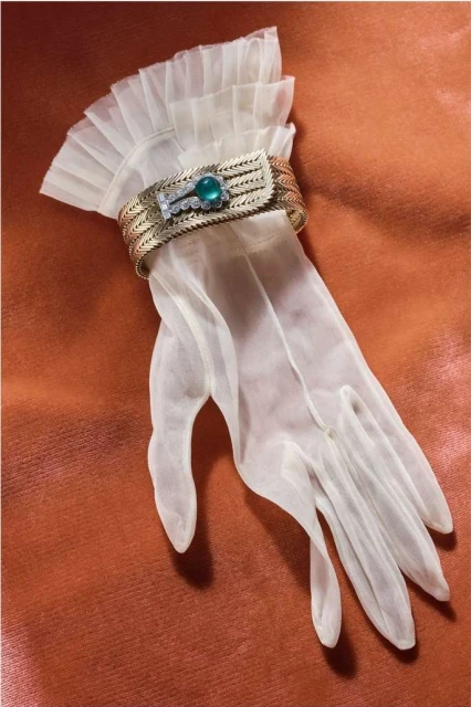 Elegant sheer glove with a ruffled cuff and a vintage gold bracelet with green gem displayed on a textured orange surface.