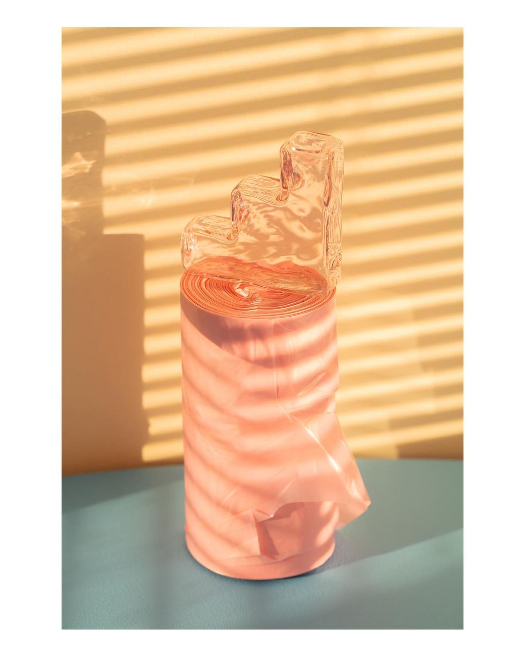 Still life featuring a roll of pink plastic bags with a clear glass sculptural block, set against a yellow and blue backdrop with shadows.