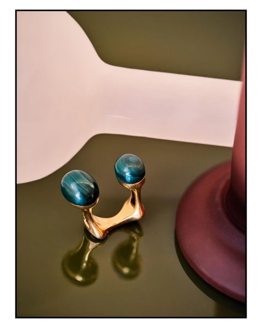 Close-up of a golden facial massage roller with teal jade stones on a glossy green surface with pink and burgundy abstract geometric shapes.