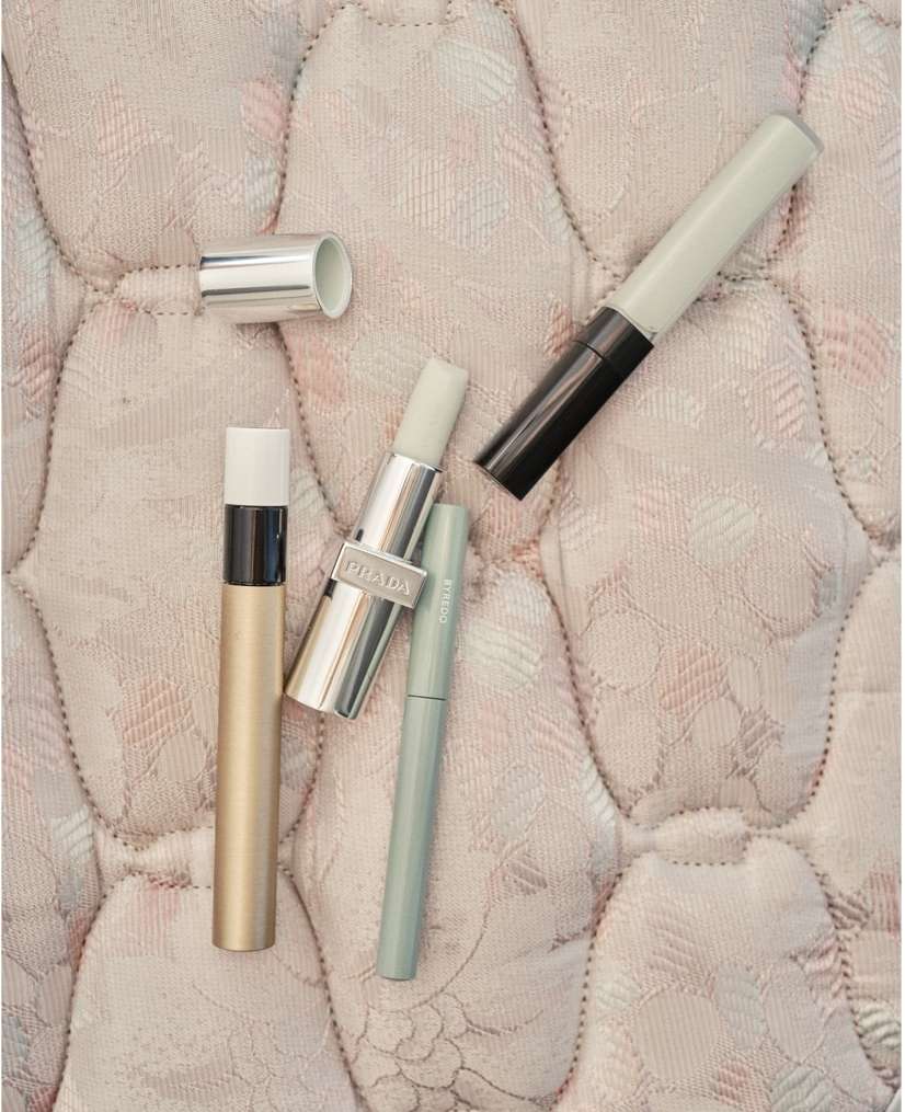 Four beauty products including lipstick and eye makeup on a quilted cream surface, showcasing cosmetic essentials in a minimalist composition.