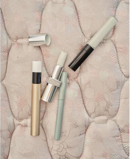 Four beauty products including lipstick and eye makeup on a quilted cream surface, showcasing cosmetic essentials in a minimalist composition.