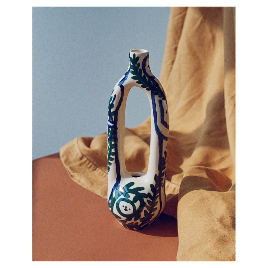 Modern ceramic vase with a hollowed center and painted with leafy patterns on a peach surface in front of a draped linen cloth.