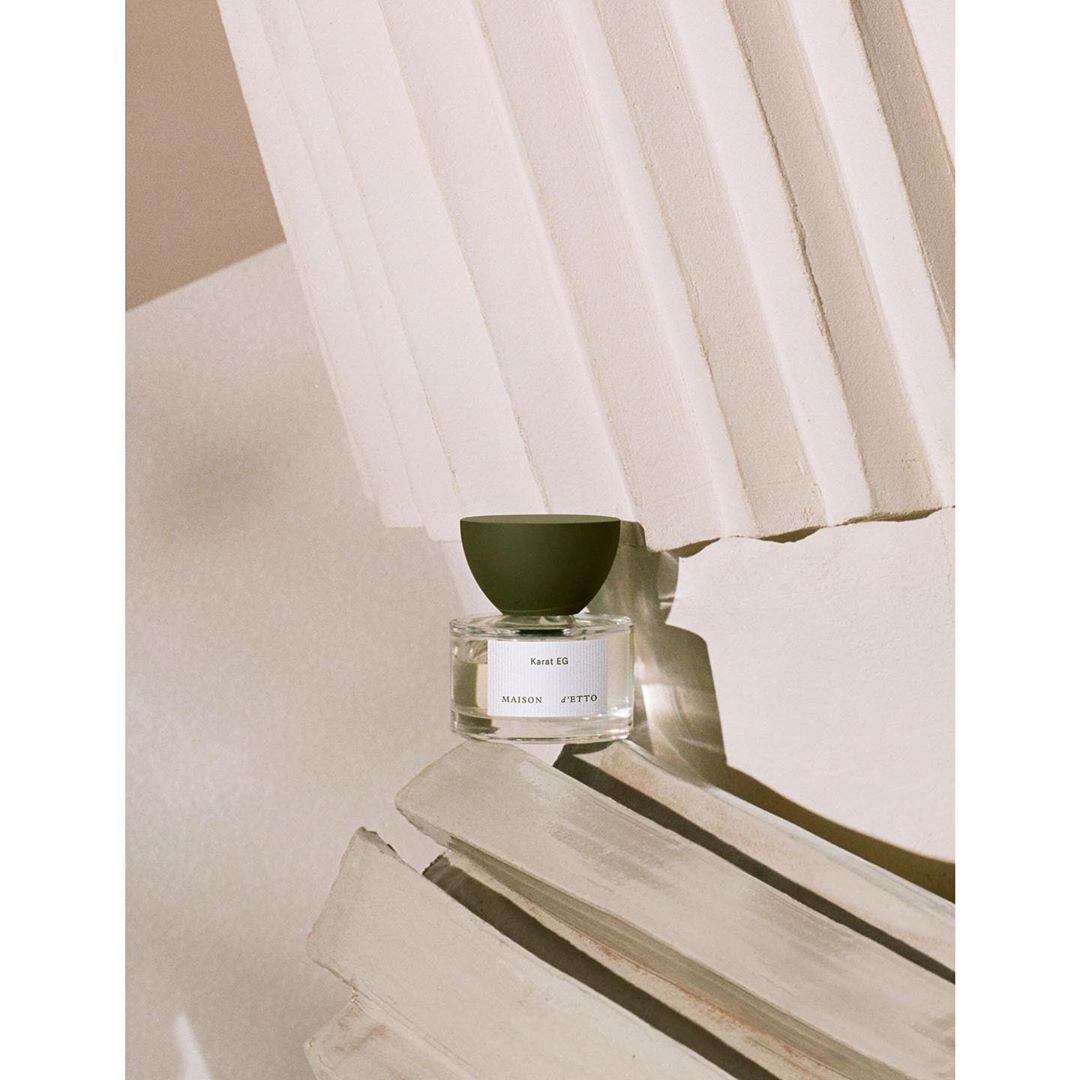 Studio shot of a bottle of Maison JYTTO fragrance on a decorative off-white surface with an olive green cap and visible text.