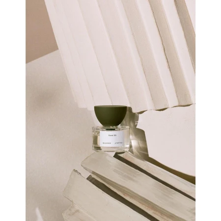 Studio shot of a bottle of Maison JYTTO fragrance on a decorative off-white surface with an olive green cap and visible text.