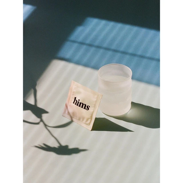 Still life of a clear glass with liquid and a Hims single-use wipe on a bright surface with blue shadows from window blinds.