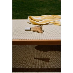 A glass bottle of rose wine and a yellow and white striped towel rest on a textured lounger outdoors.