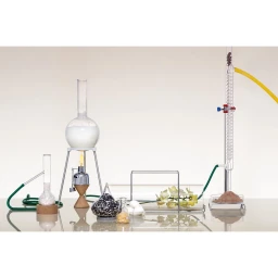 Laboratory glassware setup featuring round-bottom flasks, a condenser column, and a spirit burner on a reflective surface.
