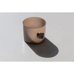 A beige glass cup with a dark stone handle rests on a smooth white surface, casting a long diagonal shadow.