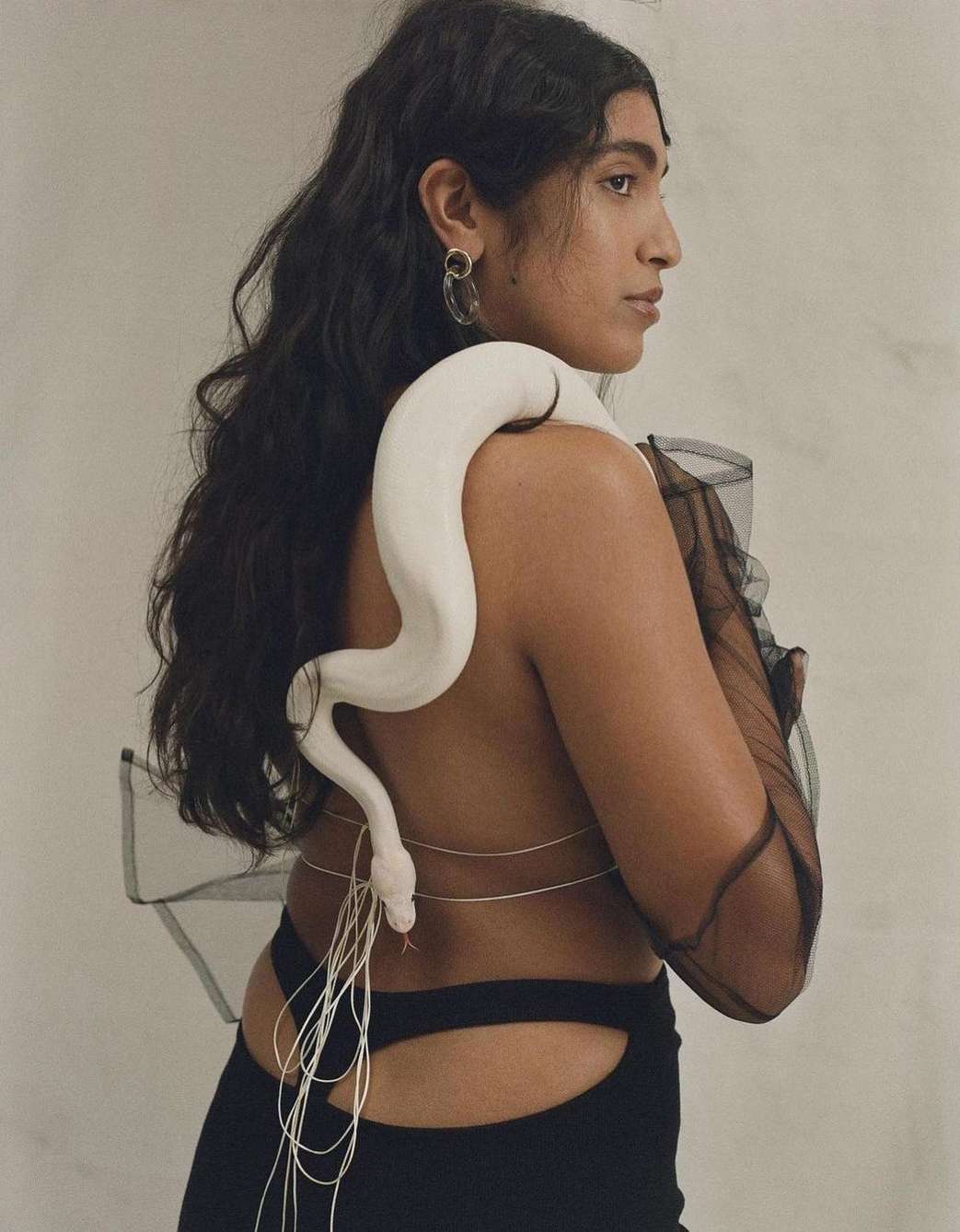 A dark-haired woman in a black dress is shown from the waist up, in a studio shot, with a large white snake draped around her shoulders.