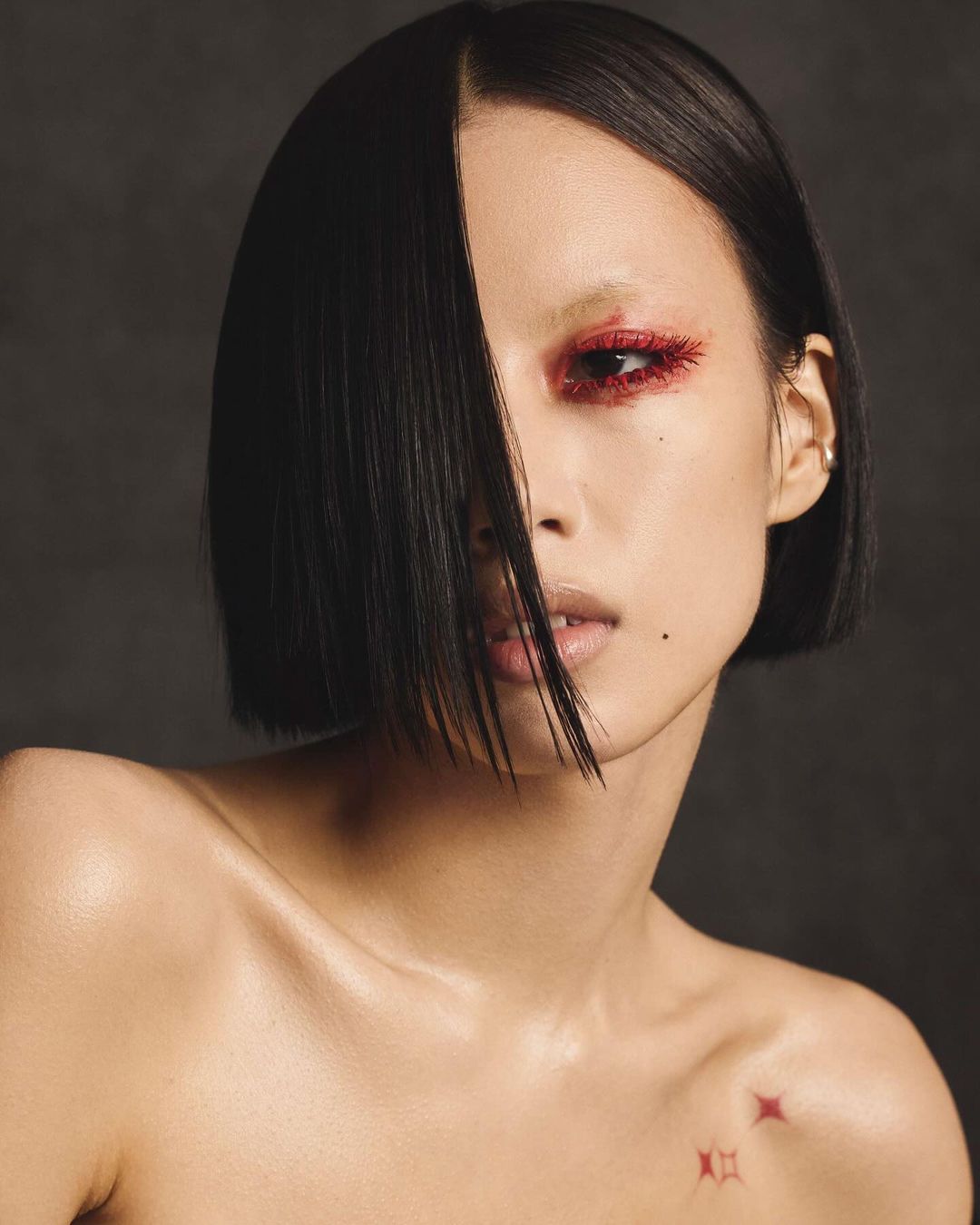 Close-up portrait of a young Asian woman with short black hair and red glittery eyeshadow against a dark background.
