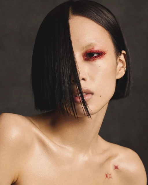 Close-up portrait of a young Asian woman with short black hair and red glittery eyeshadow against a dark background.