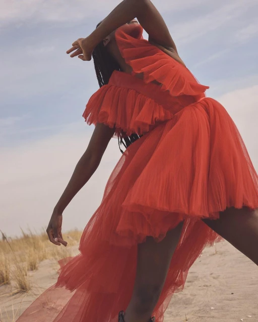 An African American woman posing on a sandy surface, wearing a bright orange, layered, ruffled dress with a blue sky in the background.