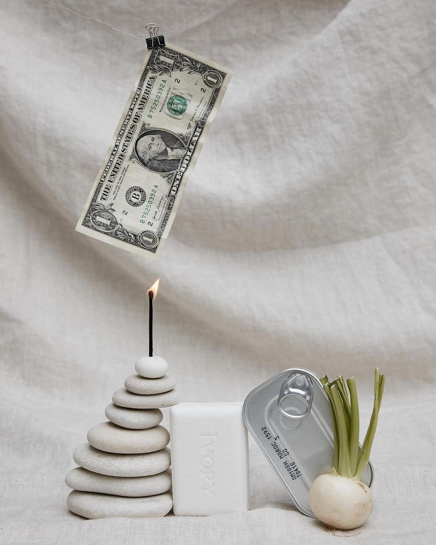 Still life photo features a one dollar bill suspended by a clip, a burning candle atop stacked stones, soap, and an open tin can with a sprout.