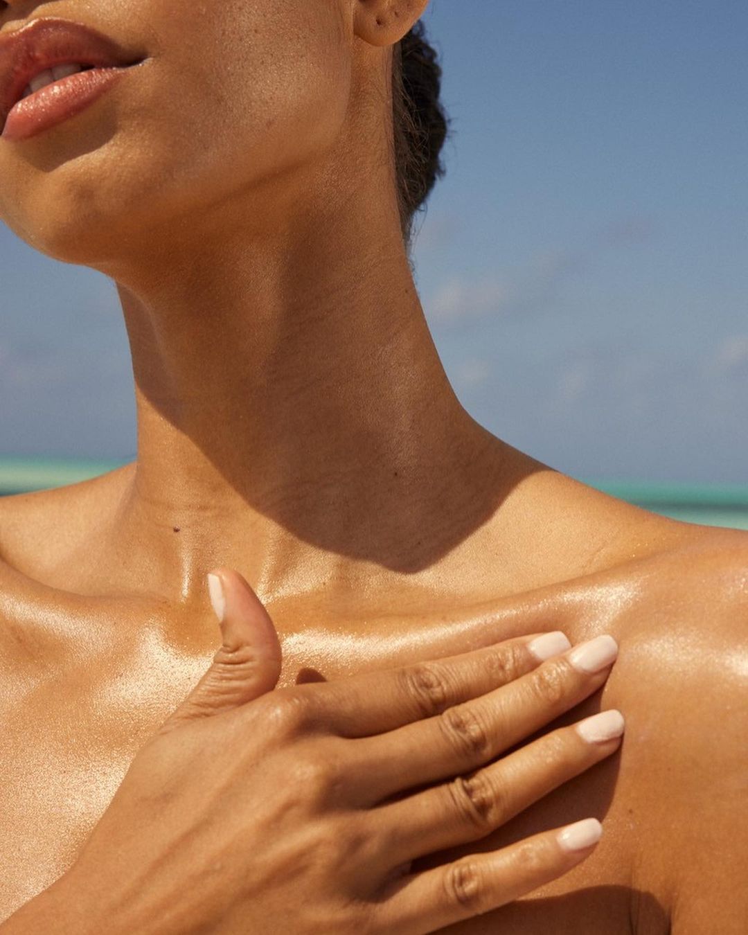 Close-up studio shot of a tanned adult woman with glossy skin and pink lipstick touching her clavicle with a manicured hand against a blue sky.