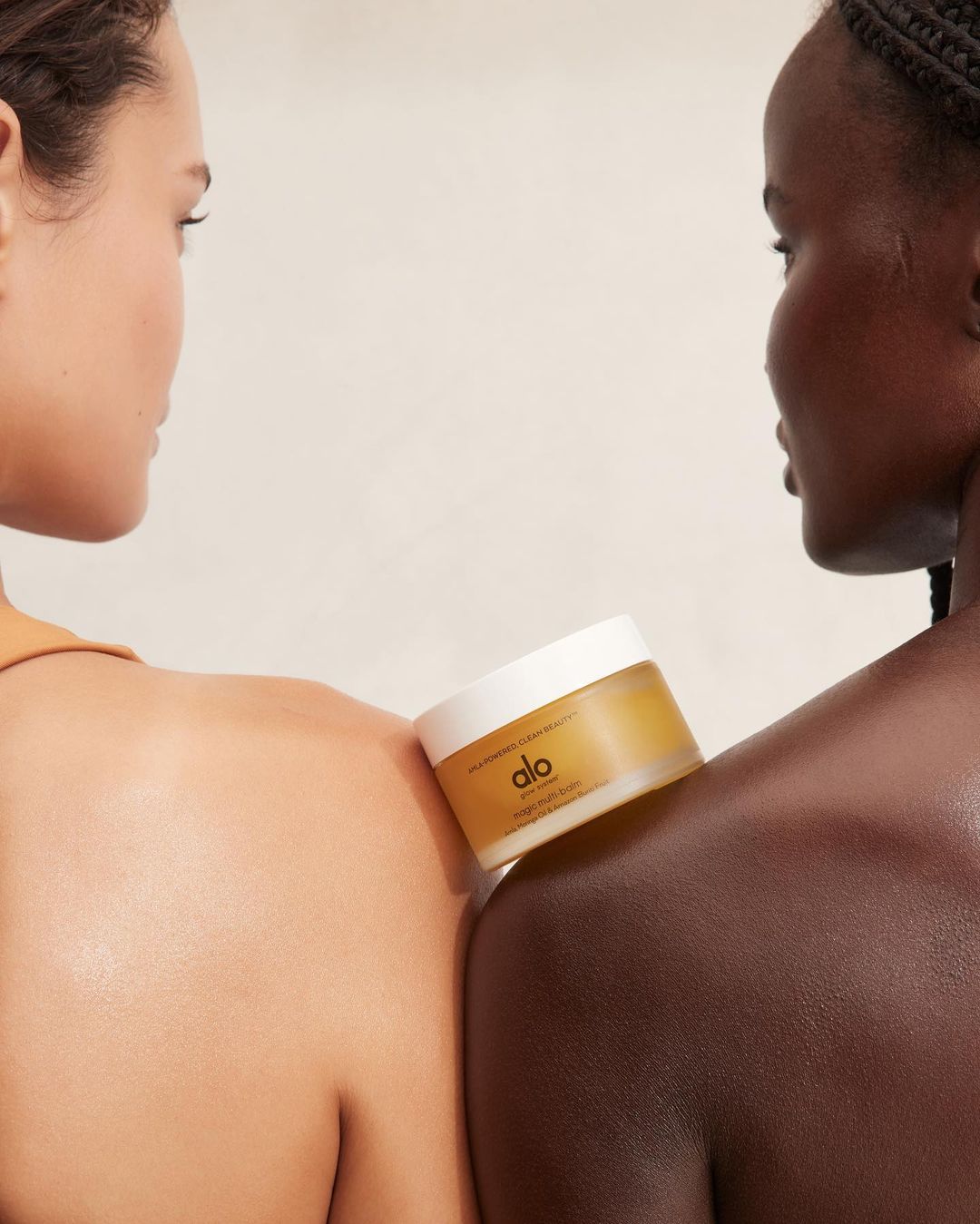 Two women of different ethnicities pose closely with Alo body balm on shoulders, showcasing beauty and skincare product in studio setting.