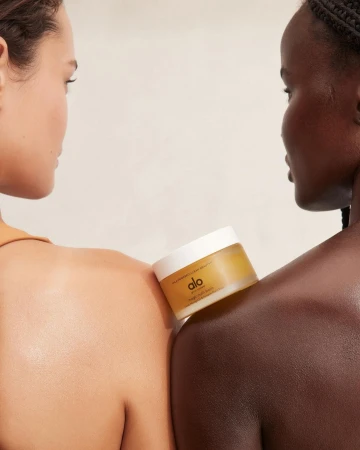 Two women of different ethnicities pose closely with Alo body balm on shoulders, showcasing beauty and skincare product in studio setting.