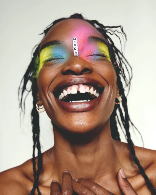 Close-up of a smiling dark-skinned woman with colorful powder makeup, braids, gold hoops, and letter beads on her forehead. 