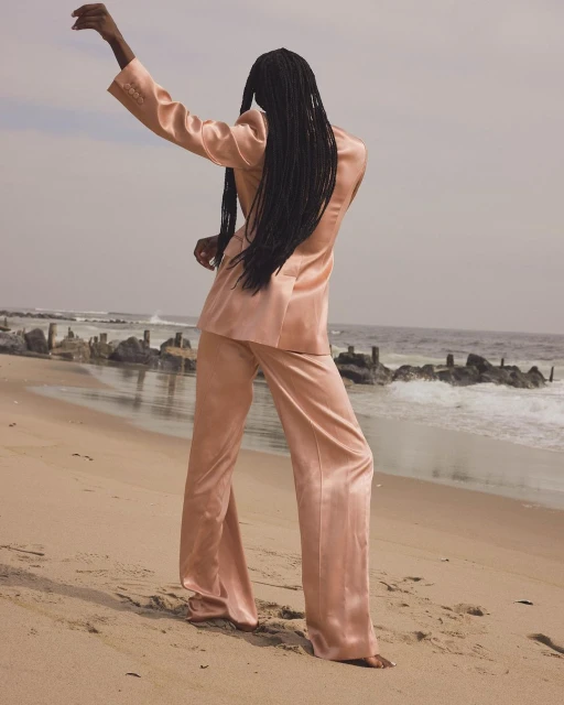 Woman in salmon-colored suit standing barefoot on beach. Ocean waves and horizon in background.