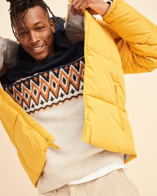 Smiling man in a yellow puffer jacket and patterned sweater poses indoors, showcasing winter fashion with a bright, joyful expression.
