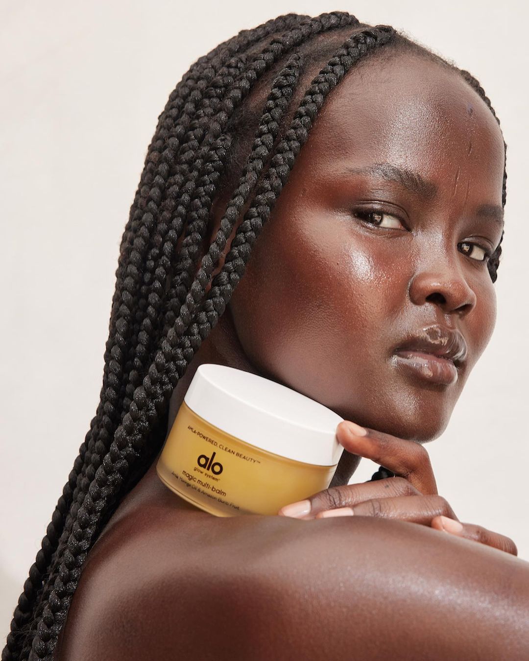 A dark-skinned woman with braided hair holds a jar of Alo moisturizer against a cream background, showcasing beauty and skincare.