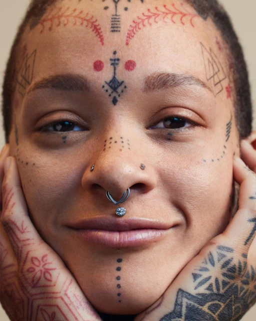 Close-up portrait of a smiling adult female with tribal-style face and hand tattoos, silver nose and lip rings, and hands framing face.