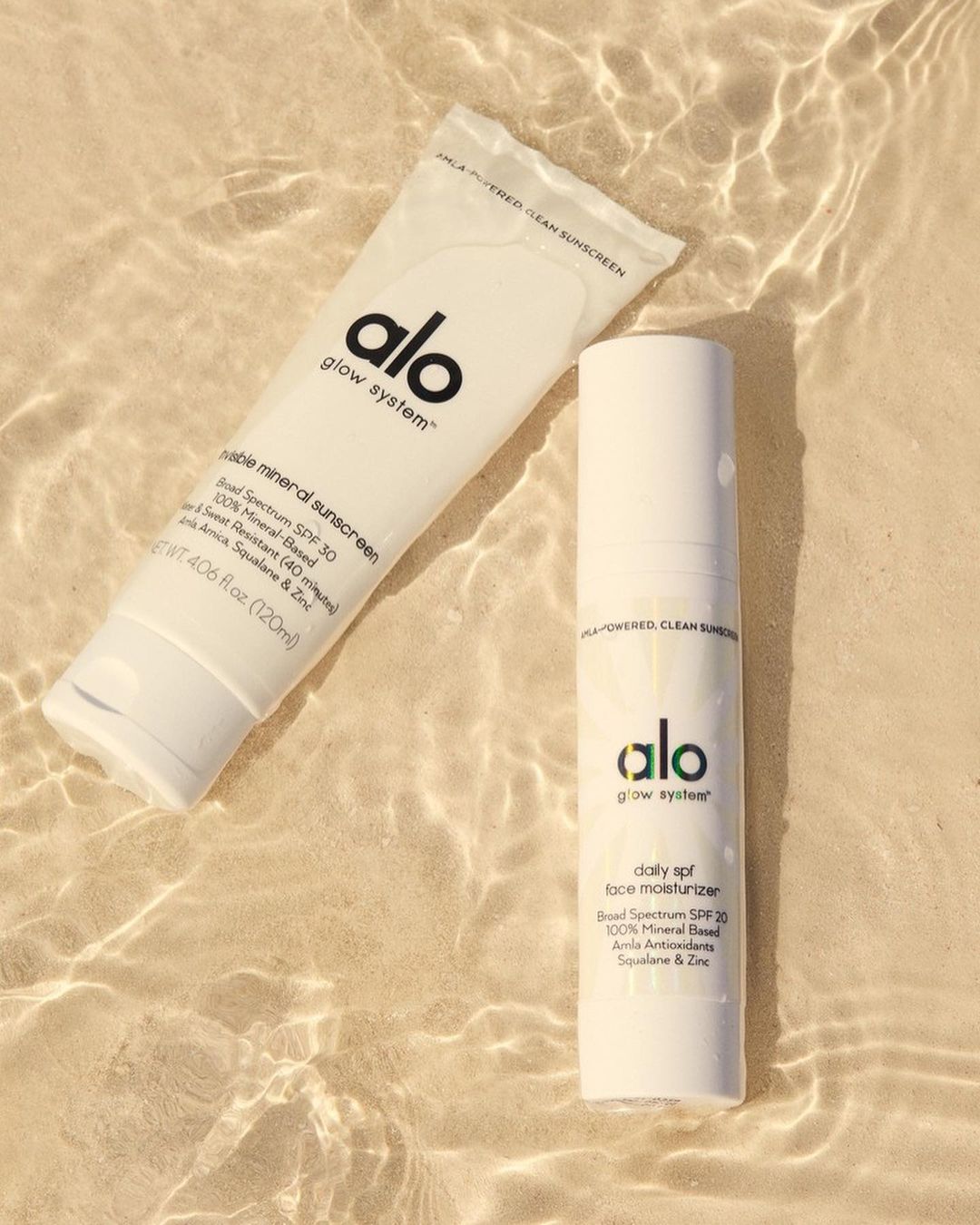 Two white bottles of Alo Glow System sunscreen and moisturizer are displayed against a beige sandy background with water reflections.