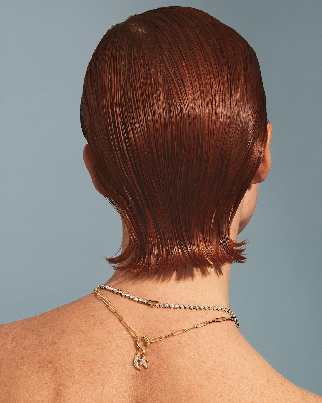 Rear view of an adult female with auburn hair wearing a gold necklace with a moon and star charm against a light blue backdrop.