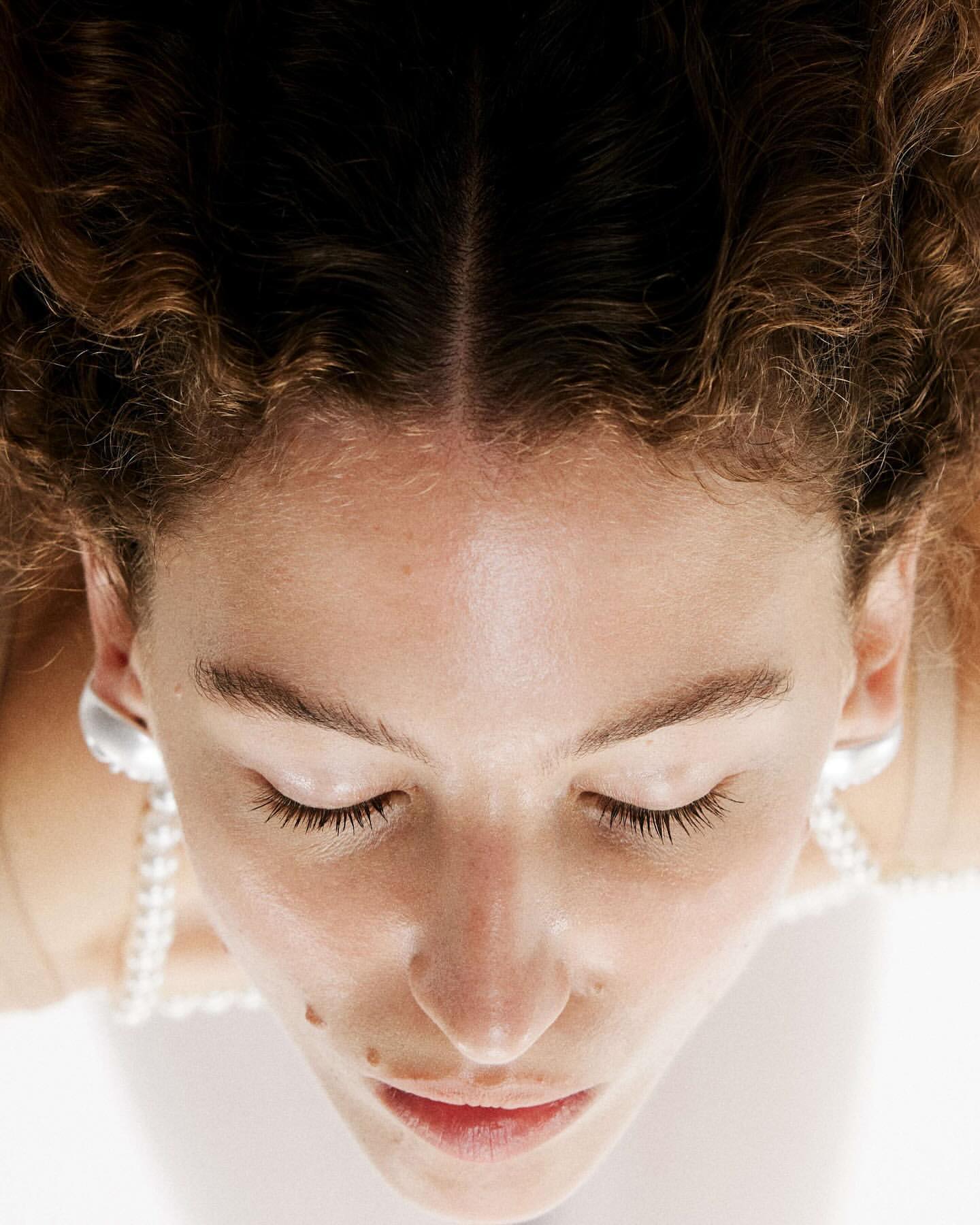 Close-up overhead portrait of a woman with curly brown hair, closed eyes, pearl necklace, and silver earrings against a white background.