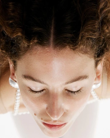 Close-up overhead portrait of a woman with curly brown hair, closed eyes, pearl necklace, and silver earrings against a white background.