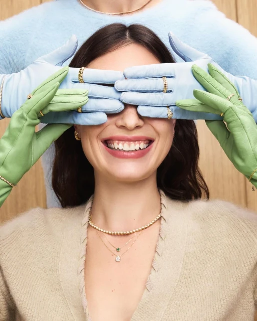 Smiling woman with hands in blue and green gloves covering eyes, wearing multiple gold necklaces and rings, and a beige sweater.