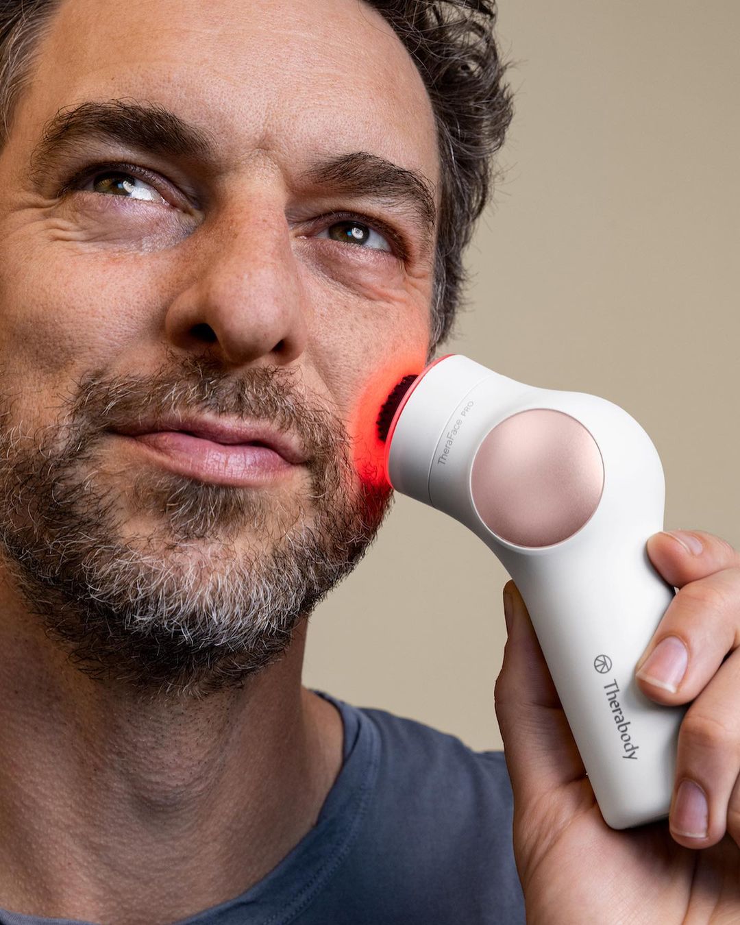 Adult man using Therabody TheraFace Pro handheld device on face with red light. Beauty and skincare treatment on neutral backdrop.