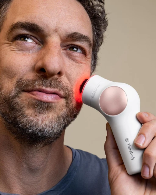 Adult man using Therabody TheraFace Pro handheld device on face with red light. Beauty and skincare treatment on neutral backdrop.
