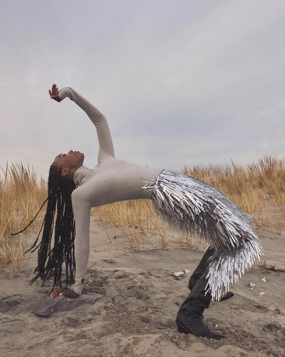 Adult female in a light beige long-sleeve top and silver fringe skirt arches her back on a sandy beach with dune grass under a cloudy sky.