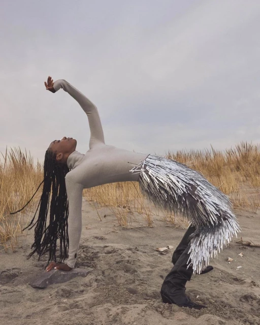 Adult female in a light beige long-sleeve top and silver fringe skirt arches her back on a sandy beach with dune grass under a cloudy sky.