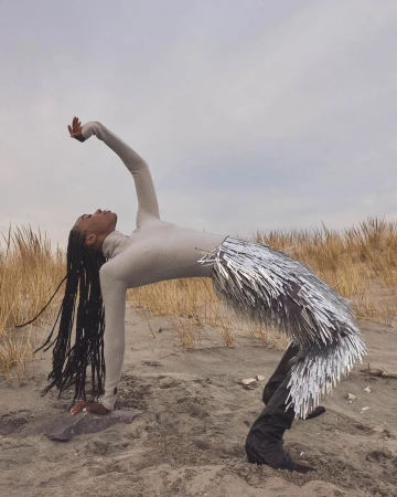 Adult female in a light beige long-sleeve top and silver fringe skirt arches her back on a sandy beach with dune grass under a cloudy sky.