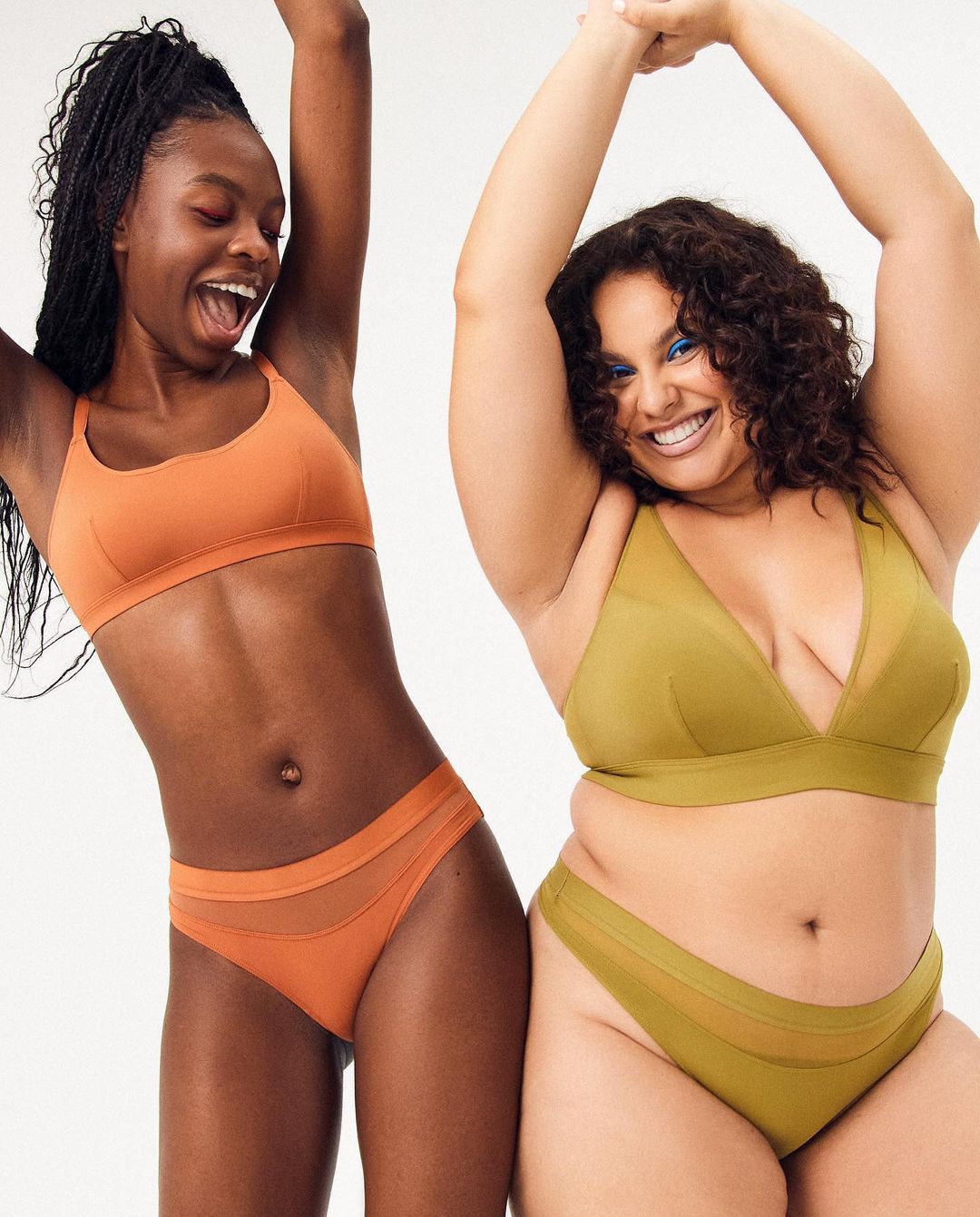 Two women joyfully pose in matching underwear sets against a bright backdrop, smiling with arms raised, showcasing body positivity and comfortable fashion.