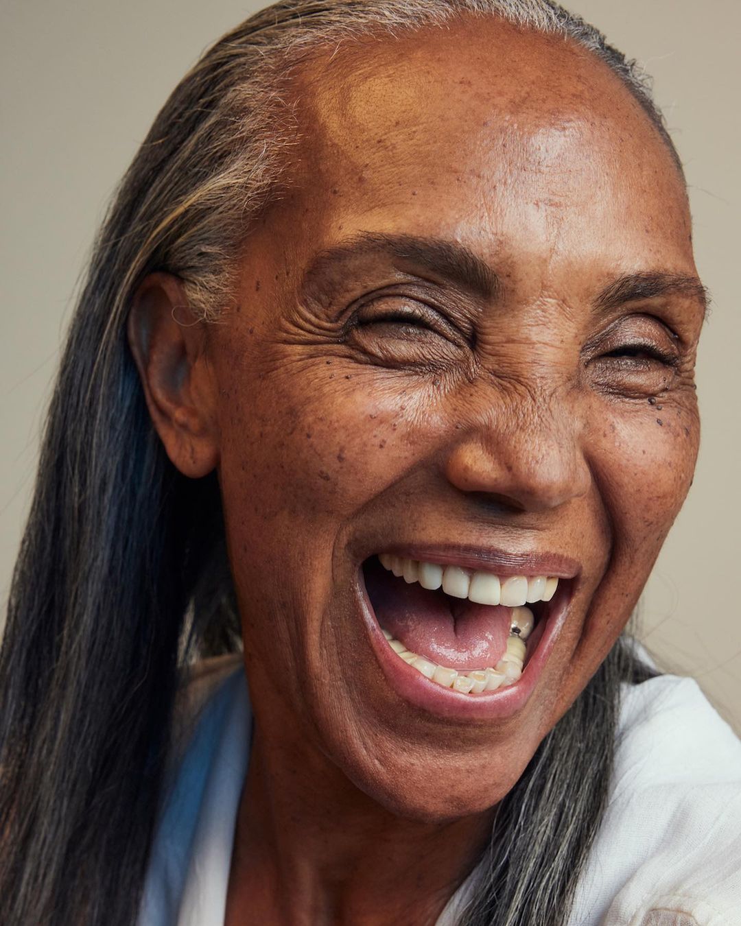 Close-up portrait of an older Black woman with graying hair laughing. Her teeth are visible. She is wearing a white shirt.