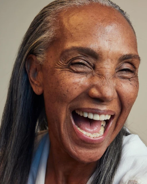 Close-up portrait of an older Black woman with graying hair laughing. Her teeth are visible. She is wearing a white shirt.