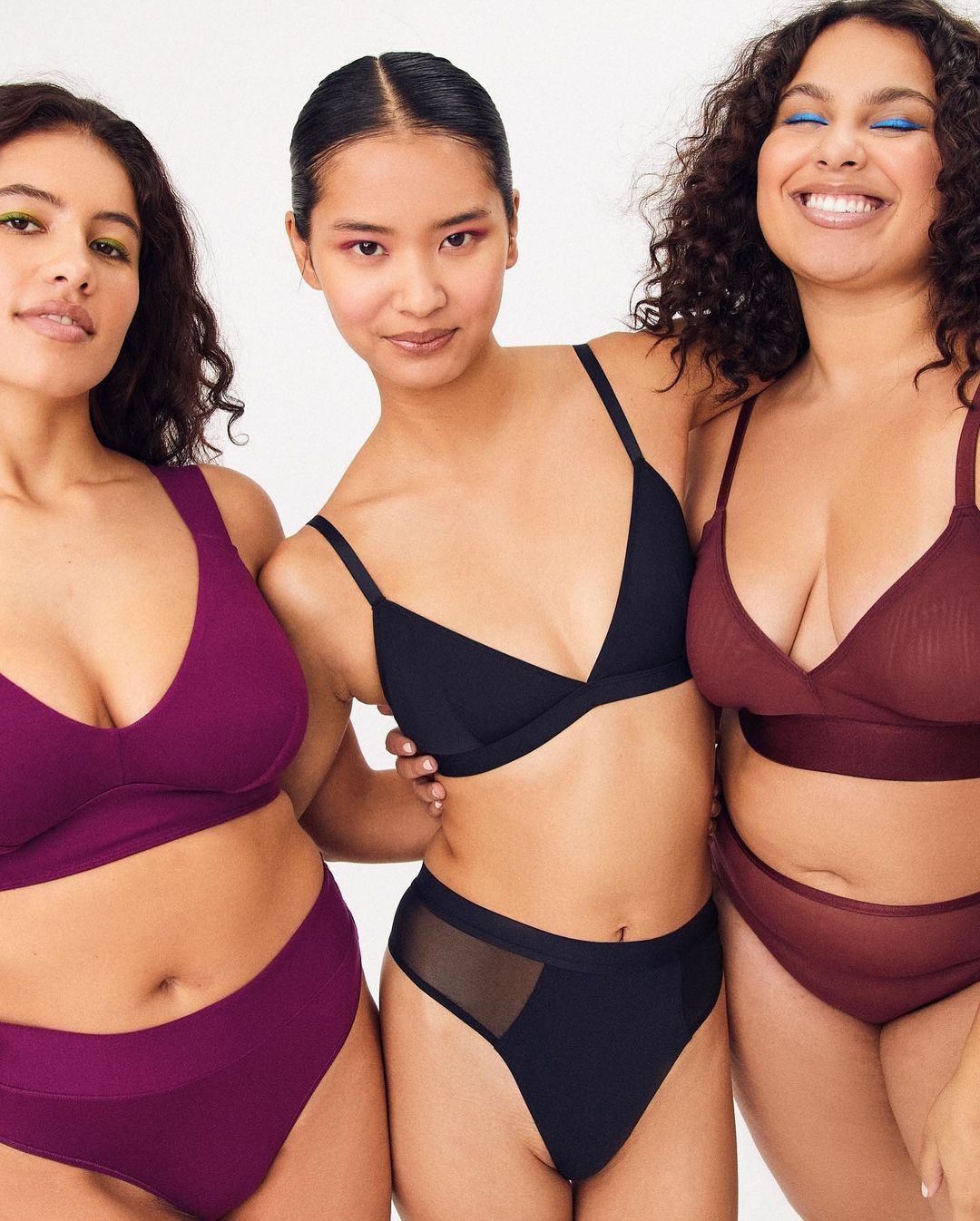Three women in matching underwear sets pose in front of a white background. The woman on the right smiles with her eyes closed.