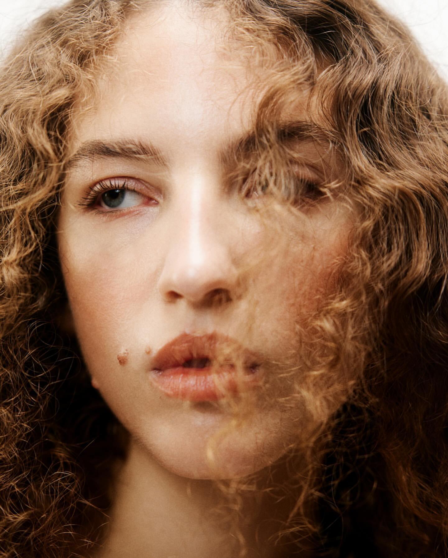 Close-up portrait of a young woman with curly blonde hair falling across her face against a neutral background.