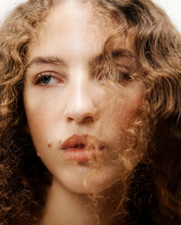 Close-up portrait of a young woman with curly blonde hair falling across her face against a neutral background.