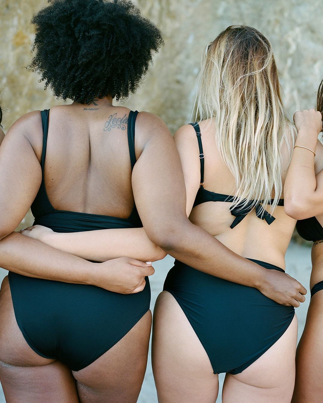 Three women in black swimsuits stand with their arms around each other in front of a neutral wall, showcasing swimwear and friendship.