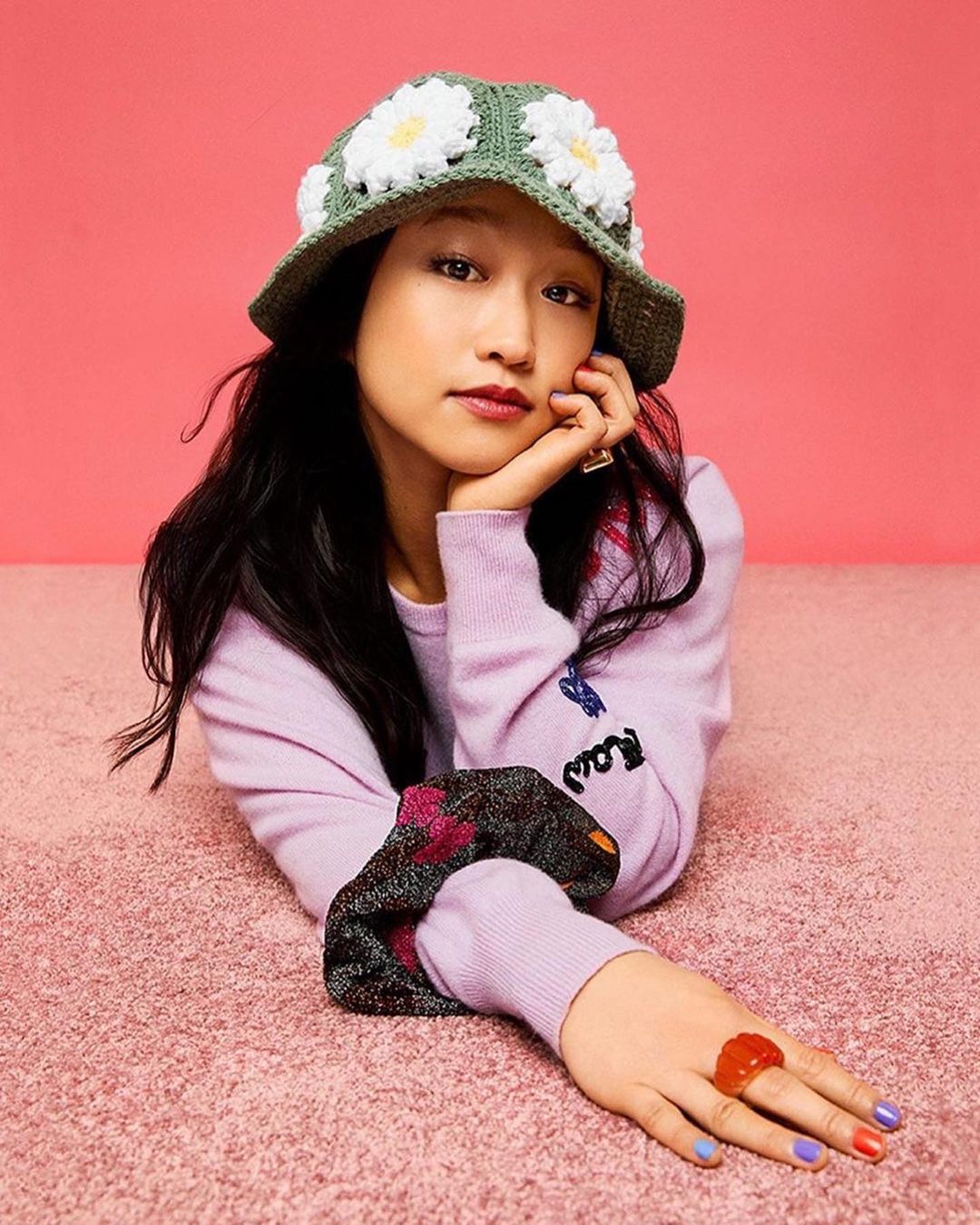 Portrait of a young Asian woman wearing a crochet hat with daisy accents, paired with rings and a lilac sweater, on a pink background.