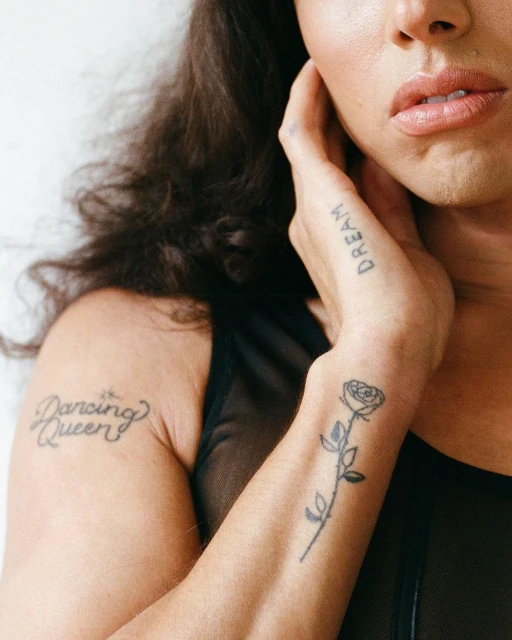 Close-up portrait of an adult female with dark hair and tattoos on her arm and hand, wearing a black mesh top against a white background.
