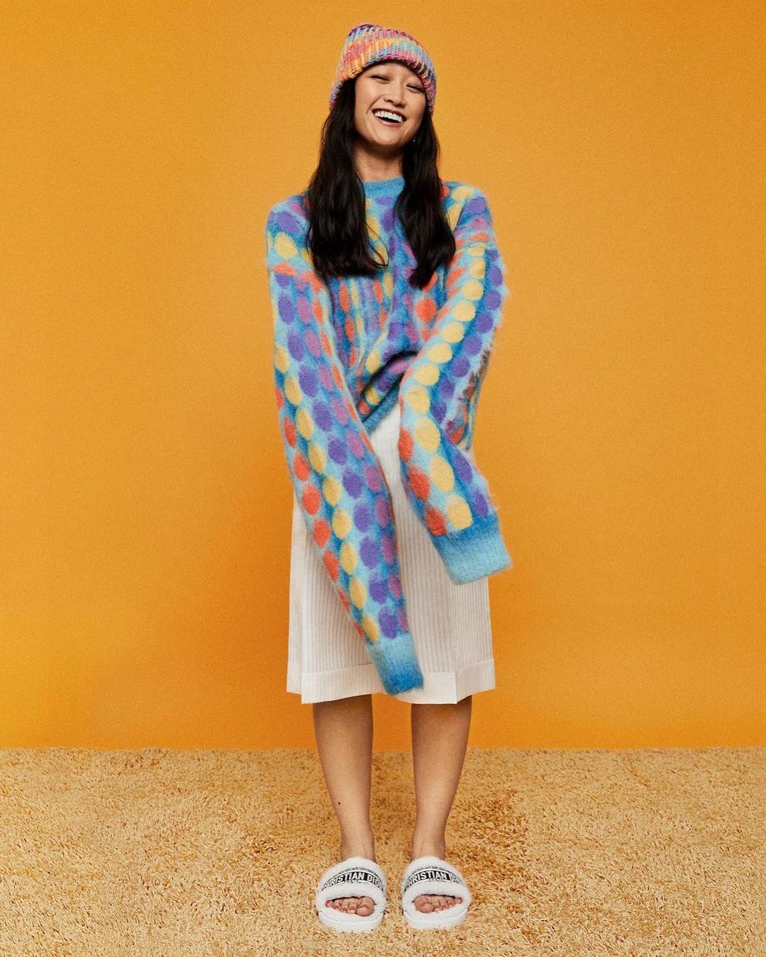 Smiling Asian woman in colorful knitwear: a beanie and sweater with polka dots, paired with a white skirt and fluffy slippers.