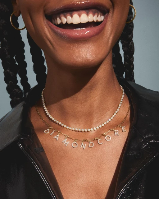 Smiling Black woman showcasing two diamond necklaces, wearing a black leather jacket against a grey background in a studio shot.