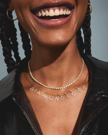 Smiling Black woman showcasing two diamond necklaces, wearing a black leather jacket against a grey background in a studio shot.